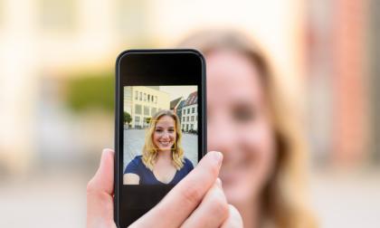 Happy attractive young blond woman taking a selfie outdoors in an urban street with ficus to her image on the screen of her mobile phone