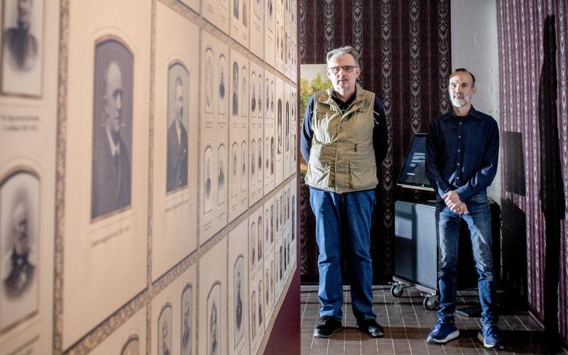 Etnolog Conny Andersson och överintendent Graham Robins framför väggen med de 90 porträtten i familjen Öhbergs album.